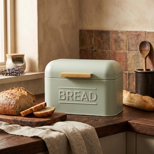 Peut inclure: Une boîte à pain vert sauge avec le mot "BREAD" en relief sur le devant. La boîte a une poignée en bambou et un dessus arrondi. Un pain et des tranches sont sur une planche à découper en bois.