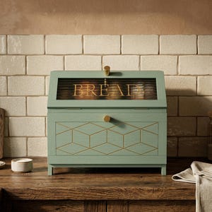 Puede incluir: Una panera verde menta con una ventana de cristal que muestra la palabra "BREAD" en letras doradas. La panera tiene herrajes dorados y un diseño geométrico en la parte delantera. Se encuentra sobre una encimera de madera.