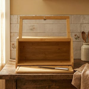 Puede incluir: Una panera de madera con una puerta de cristal transparente y acanalado. La panera está abierta, revelando una tabla de cortar y un cuchillo. La panera es de madera clara y está sobre una mesa de madera. El fondo incluye una pared de baldosas de cerámica.