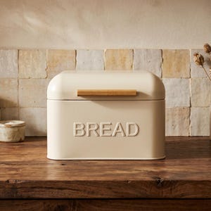 May include: Cream-coloured bread bin with a rounded top and a wooden handle. The word "BREAD" is embossed on the front. The bin is made of metal and sits on a wooden surface.