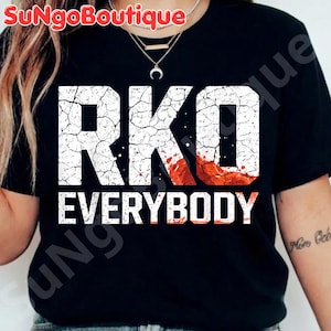 Puede incluir: Camiseta negra con el texto blanco "RKO EVERYBODY". Las letras "RKO" tienen una textura agrietada, con un efecto de salpicadura rojo y naranja. La camiseta es de cuello redondo.