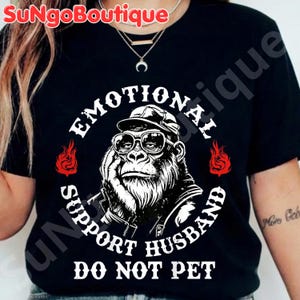May include: Black t-shirt featuring a gorilla wearing glasses and a cap, with the text "EMOTIONAL SUPPORT HUSBAND DO NOT PET" in a circular design. Red flame graphics flank the gorilla image. The brand name "SuNGoBoutique" is at the top.