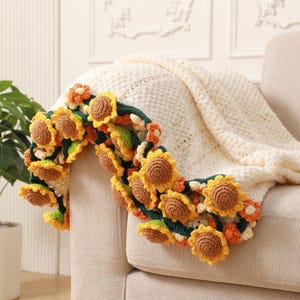 Peut inclure: Une guirlande de tournesols crochetée drapée sur une couverture crème sur un canapé beige. La guirlande présente des tournesols jaunes, bruns et verts avec des fleurs d'accent orange et blanches. La couverture a un motif texturé.
