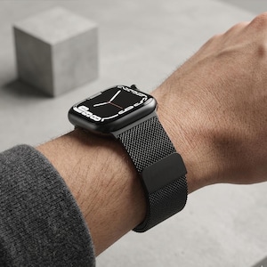 May include: A black smartwatch with a black mesh band is displayed on a wrist. The watch face is black with white numerals and hands. The watch is set against a gray background with a gray cube.