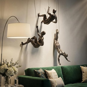 Könnte beinhalten: Drei metallische, bronzefarbene Wandskulpturen, die Figuren in Kletter- und Abseilposen darstellen. Jede Figur ist an einem dünnen, an der Wand befestigten Draht aufgehängt, was eine Illusion von Bewegung auf der vertikalen Fläche erzeugt.