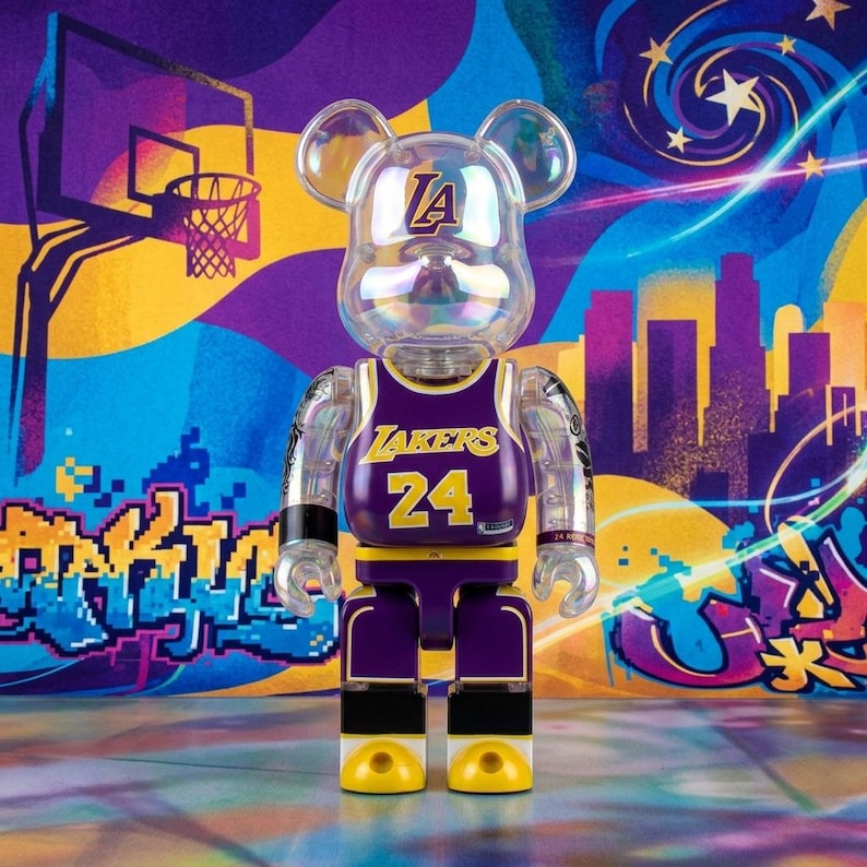 Op de afbeelding: Een doorzichtig, beer-vormig verzamelfiguur dat een paars en goud basketbalshirt van de Los Angeles Lakers draagt met het nummer 24 op de voorkant. Het figuur heeft een Lakers-logo op het voorhoofd en zwarte polsbandjes om beide armen.