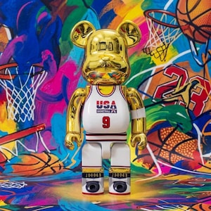 Pu&ograve; includere: Una statuetta da collezione a forma di orso color oro che indossa una maglia da basket USA bianca con il numero 9 stampato sul davanti. La figura presenta una finitura metallica riflettente, pantaloncini bianchi con bordi neri e rossi e scarpe nere con la scritta JORDAN.