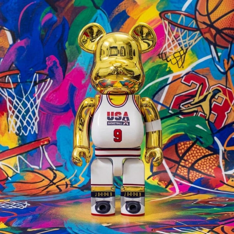 Pu&ograve; includere: Una statuetta da collezione a forma di orso color oro che indossa una maglia da basket USA bianca con il numero 9 stampato sul davanti. La figura presenta una finitura metallica riflettente, pantaloncini bianchi con bordi neri e rossi e scarpe nere con la scritta JORDAN.