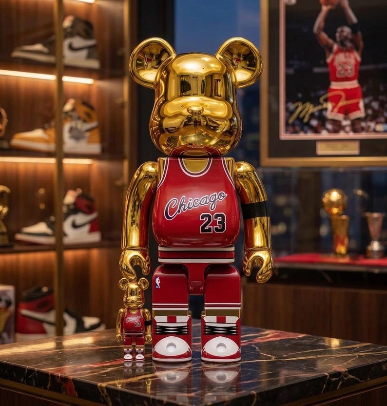 K&ouml;nnte beinhalten: Eine gro&szlig;e und eine kleine goldene b&auml;renf&ouml;rmige Sammlerfigur, die ein rotes Chicago 23 Basketballtrikot tr&auml;gt. Die Figuren haben eine reflektierende metallische Oberfl&auml;che, wobei das Trikot wei&szlig;e R&auml;nder, den Schriftzug Chicago und die Nummer 23 in Schwarz aufweist.