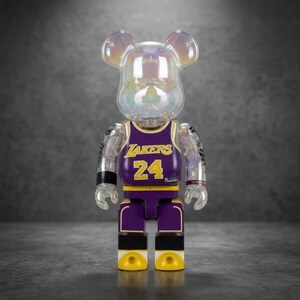 Op de afbeelding: Een doorschijnend iriserend beer-vormig verzamelfiguur dat een paars en geel basketbalshirt draagt. Het shirt is voorzien van de tekst LAKERS en het nummer 24 in het geel. Het figuur heeft beweegbare ledematen en een ronde kop met oren.