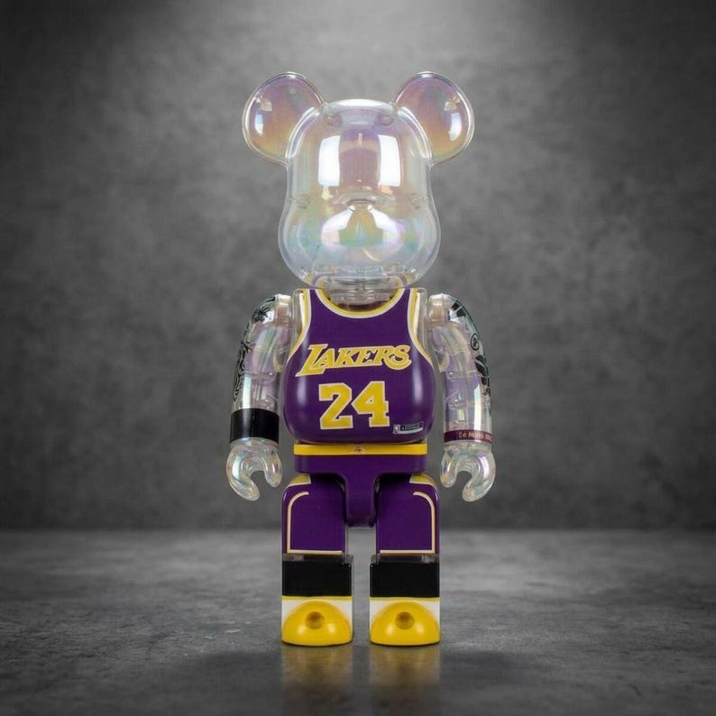 Op de afbeelding: Een doorschijnend iriserend beer-vormig verzamelfiguur dat een paars en geel basketbalshirt draagt. Het shirt is voorzien van de tekst LAKERS en het nummer 24 in het geel. Het figuur heeft beweegbare ledematen en een ronde kop met oren.