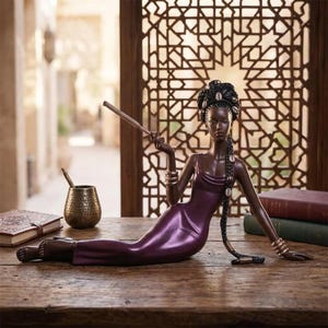 May include: A dark-skinned figurine of a woman in a purple dress, reclining on a wooden surface. The figure holds a long, thin object and is adorned with bracelets and seashell hair accessories. The background features a decorative wooden screen.