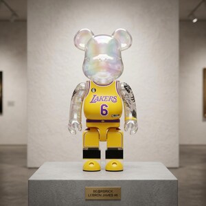 Pu&ograve; includere: Una statuetta Bearbrick trasparente e iridescente che indossa una maglia gialla dei Los Angeles Lakers con il numero 6. La figura presenta tatuaggi neri sulle braccia, una testa a forma di orso e una targa BE@RBRICK LEBRON JAMES #6.