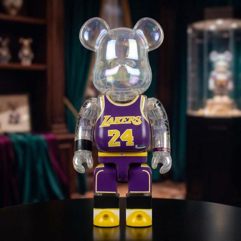 Op de afbeelding: Een doorzichtig, iriserend, beer-vormig verzamelfiguur dat een paars en geel basketbalshirt draagt. Het shirt is voorzien van de tekst LAKERS en het nummer 24 in het geel. Het figuur heeft beweegbare ledematen, een ronde kop en oren.