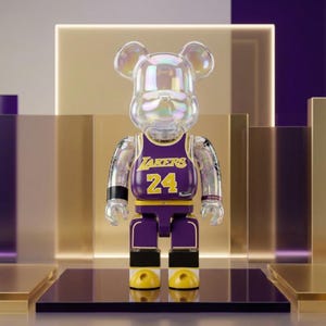 Op de afbeelding: Een verzamelbaar beer-vormig figuur met een doorschijnend, iriserend hoofd en ledematen. Het draagt een paars en geel basketbalshirt met de tekst LAKERS en het nummer 24 op de borst. Het figuur heeft zwarte polsbanden en gele voeten.