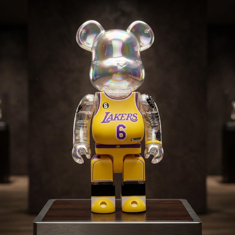 Pu&ograve; includere: Una statuetta da collezione a forma di orso con testa e arti trasparenti e iridescenti. Il busto presenta il design di una maglia da basket gialla dei Los Angeles Lakers con la scritta Lakers e il numero 6 in viola, oltre a un piccolo numero 6 sul petto.