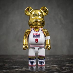 Pu&ograve; includere: Una statuetta da collezione a forma di orso placcata in oro che indossa una canotta da basket bianca con la scritta USA BASKETBALL e il numero 9 in rosso. La figura ha braccia e orecchie dorate, con gambe bianche che riportano la scritta JORDAN e dettagli blu.