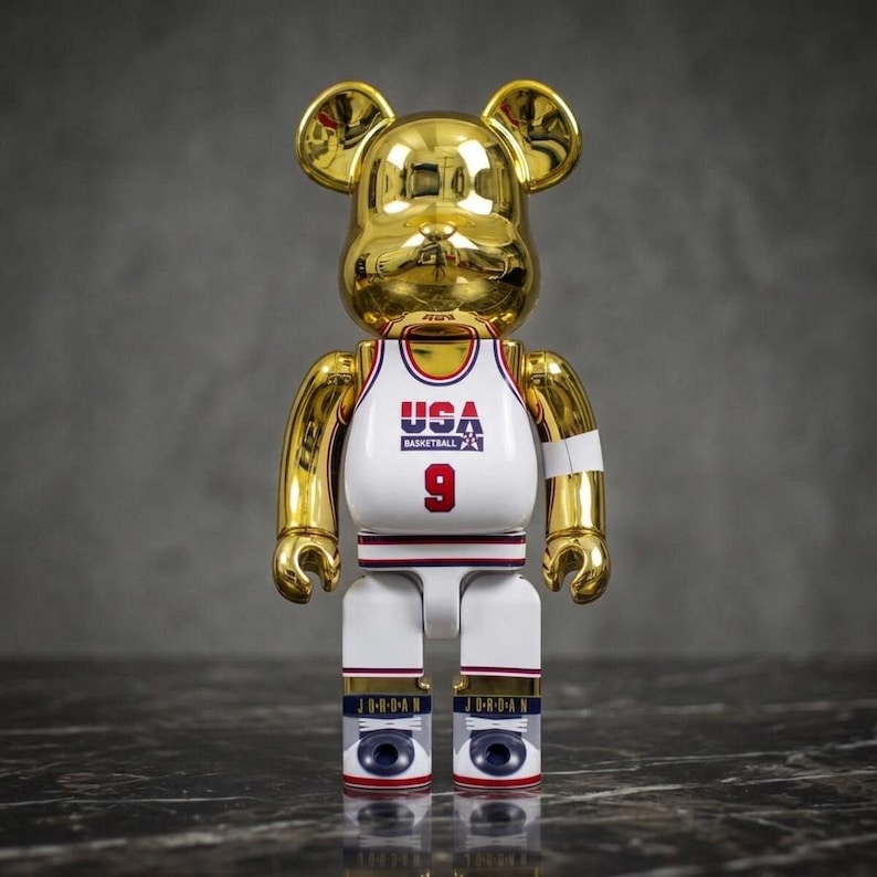 Pu&ograve; includere: Una statuetta da collezione a forma di orso placcata in oro che indossa una canotta da basket bianca con la scritta USA BASKETBALL e il numero 9 in rosso. La figura ha braccia e orecchie dorate, con gambe bianche che riportano la scritta JORDAN e dettagli blu.