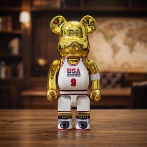 Pu&ograve; includere: Una statuetta da collezione a forma di orso di colore dorato con finitura riflettente. La figura indossa una canotta da basket bianca con la scritta USA BASKETBALL e il numero 9 in rosso. Sulla parte inferiore delle gambe appare JORDAN.