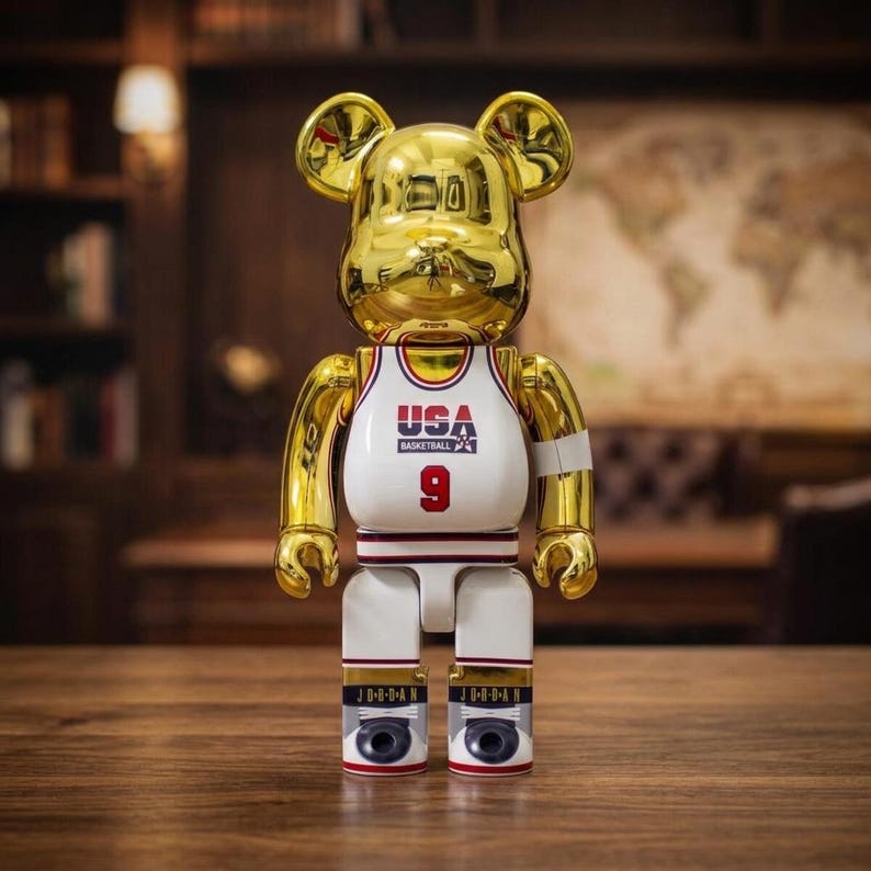 Pu&ograve; includere: Una statuetta da collezione a forma di orso di colore dorato con finitura riflettente. La figura indossa una canotta da basket bianca con la scritta USA BASKETBALL e il numero 9 in rosso. Sulla parte inferiore delle gambe appare JORDAN.
