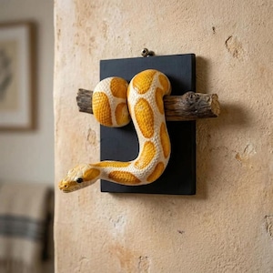 May include: A wall-mounted display featuring a yellow and white patterned snake figure coiled around a horizontal brown textured branch. The branch is attached to a rectangular black wooden plaque, which is secured to the wall with a single screw.
