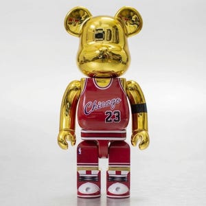 K&ouml;nnte beinhalten: Eine goldfarbene, b&auml;renf&ouml;rmige Sammlerfigur mit reflektierender Oberfl&auml;che. Die Figur tr&auml;gt ein rotes Basketballtrikot mit dem Schriftzug Chicago und der Nummer 23 in Wei&szlig; mit blauen Umrandungen. Sie hat ein schwarzes Armband und wei&szlig;e Turnschuhe.
