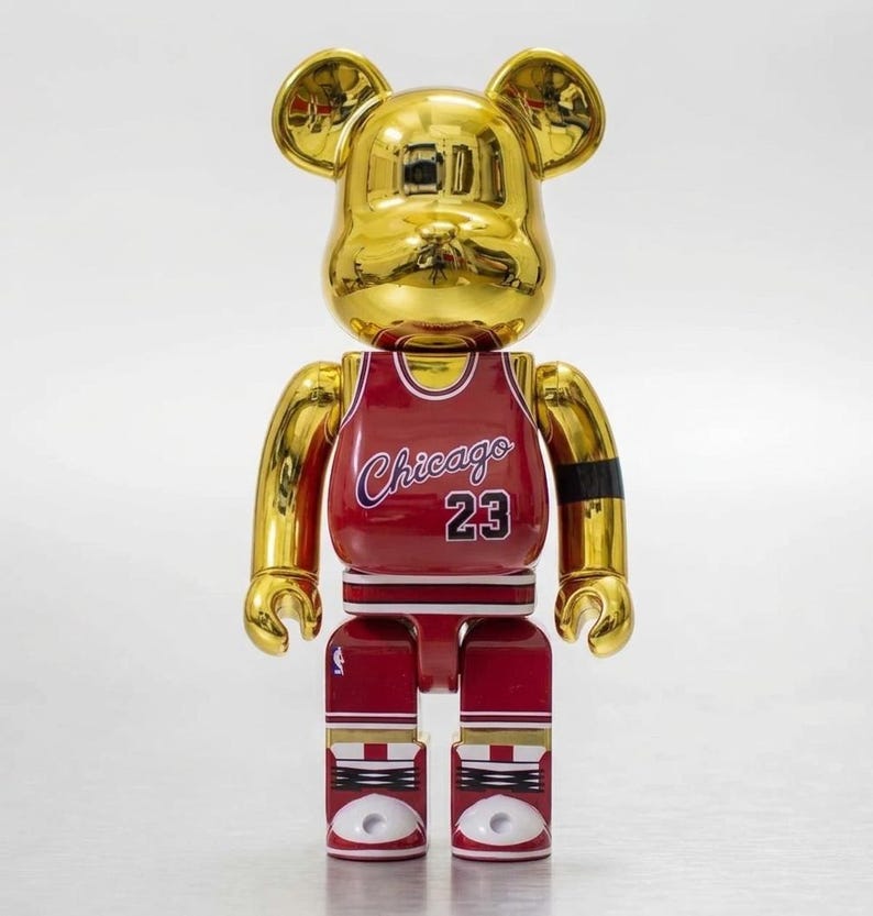 K&ouml;nnte beinhalten: Eine goldfarbene, b&auml;renf&ouml;rmige Sammlerfigur mit reflektierender Oberfl&auml;che. Die Figur tr&auml;gt ein rotes Basketballtrikot mit dem Schriftzug Chicago und der Nummer 23 in Wei&szlig; mit blauen Umrandungen. Sie hat ein schwarzes Armband und wei&szlig;e Turnschuhe.