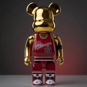 K&ouml;nnte beinhalten: Eine goldfarbene, b&auml;renf&ouml;rmige Sammlerfigur mit reflektierender Oberfl&auml;che. Die Figur tr&auml;gt ein rotes Basketballtrikot mit dem Schriftzug Chicago und der Nummer 23 in Wei&szlig; und Schwarz. Der untere Teil zeigt rot-wei&szlig;e Basketballschuhe.