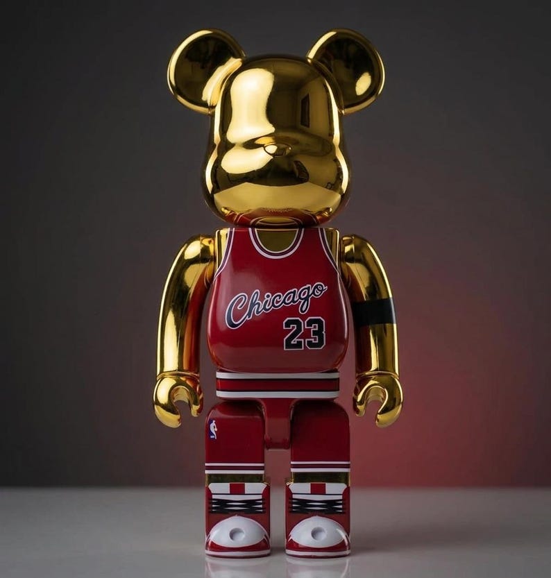 K&ouml;nnte beinhalten: Eine goldfarbene, b&auml;renf&ouml;rmige Sammlerfigur mit reflektierender Oberfl&auml;che. Die Figur tr&auml;gt ein rotes Basketballtrikot mit dem Schriftzug Chicago und der Nummer 23 in Wei&szlig; und Schwarz. Der untere Teil zeigt rot-wei&szlig;e Basketballschuhe.