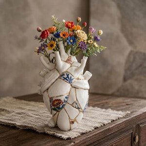 May include: A white ceramic vase shaped like an anatomical heart, featuring a kintsugi-style design with gold-filled cracks and fragments of blue and red floral patterns. The vase holds a vibrant bouquet of small, colorful artificial flowers.