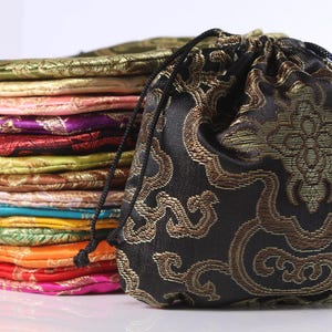 Könnte beinhalten: Ein Stapel kleiner Beutel mit Kordelzug in verschiedenen Farben, darunter Schwarz, Gold, Pink, Lila, Rot, Gelb, Blau, Grün und Orange. Jeder Beutel verfügt über ein gewebtes metallisches Muster und schwarze Kordeln zum Verschließen.
