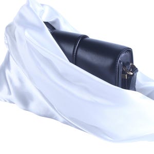 Pode incluir: Uma bolsa de couro preta com fecho dourado está parcialmente envolta em tecido de cetim branco fluido. A bolsa tem formato retangular com a parte superior arredondada e uma alça de ombro.