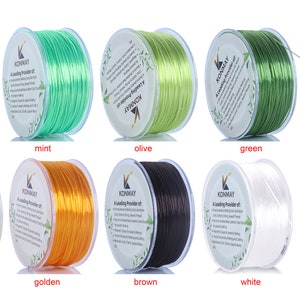 Könnte beinhalten: Zehn Spulen elastischer Schnur in verschiedenen Farben, darunter Neon-Gelb, Mint, Oliv, Grün, Dunkelgrün, Gelb, Gold, Braun, Weiß und Schwarz. Jede Spule ist mit "KONMAY" beschriftet.