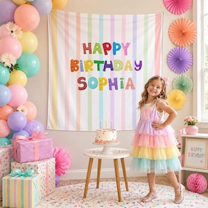 May include: A birthday party backdrop featuring vertical pastel stripes and the text HAPPY BIRTHDAY SOPHIA in colorful patterned letters. The scene includes a white cake with candles on a wooden table, wrapped gifts, and pastel balloons.