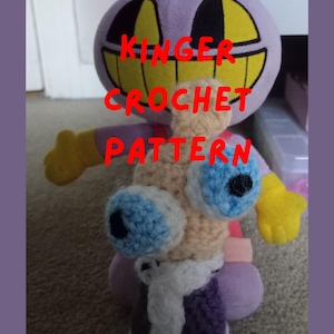 May include: A plush toy with a purple head, yellow eyes, and a wide grin. The toy has blue and white crochet eyes, yellow hands, and a purple body. Text on the image reads "Kinger Crochet Pattern PDF File."