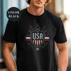 A man wearing a black t-shirt with a graphic design featuring the text "USA" and a shield-shaped emblem with stars and a soccer ball.