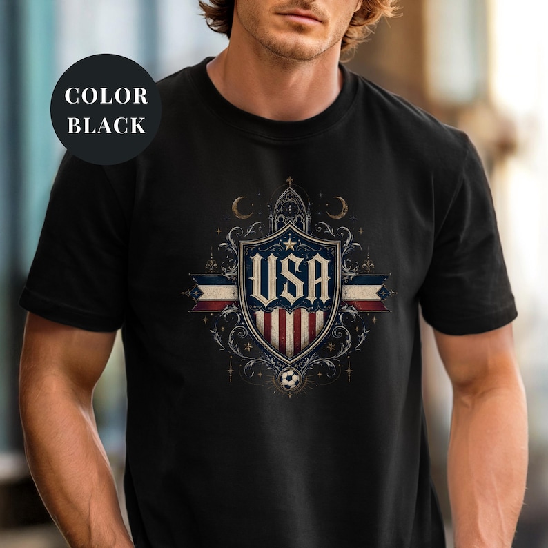 A man wearing a black t-shirt with a graphic design featuring the text "USA" and a shield-shaped emblem with stars and a soccer ball.