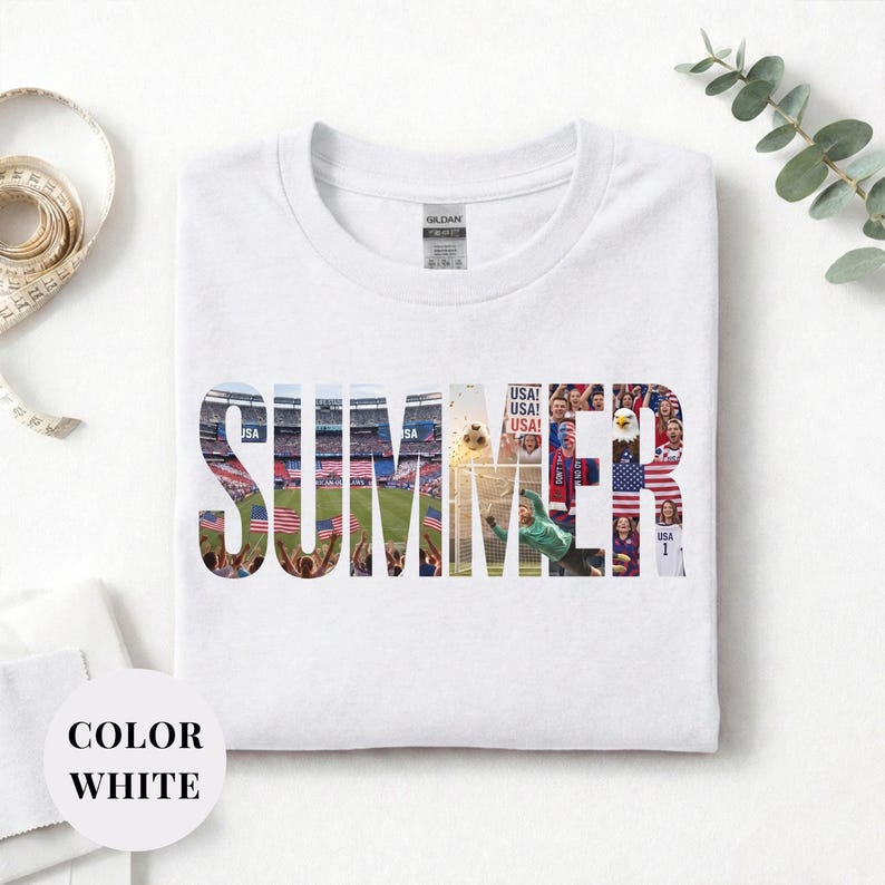 White Gildan t-shirt featuring a "SUMMER" photo collage graphic. The bold block letters are filled with USA soccer scenes, including a packed stadium, American flags, cheering fans, and a diving goalie. Patriotic sports apparel for World Cup 2026 supporters.