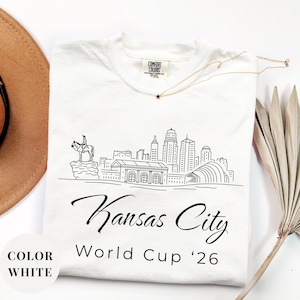 White Comfort Colors T-shirt featuring a minimalist black line art Kansas City skyline design, elegant Kansas City script lettering, and World Cup ’26 text, styled as a soccer travel souvenir shirt for Kansas City fans, football lovers, and 2026 World Cup city apparel.