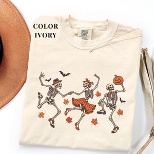 May include: An ivory t-shirt featuring a graphic of three skeletons dancing. The skeletons wear orange and black accessories, including a hat, bow tie, and polka-dot skirt. The design includes orange maple leaves, bats, and a jack-o'-lantern.