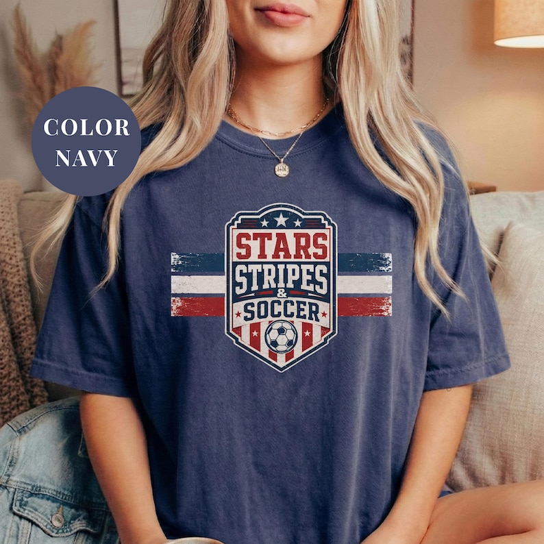 A woman wearing a navy blue t-shirt with a graphic design featuring a soccer ball and the text "STARS STRIPES & SOCCER" printed on it.