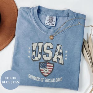 Blue Comfort Colors 1717 t-shirt with a vintage "USA Summer of Soccer 2026" graphic. Features bold varsity "USA" lettering with soccer ball patterns and a distressed American flag soccer shield. Patriotic apparel for World Cup 2026 fans.