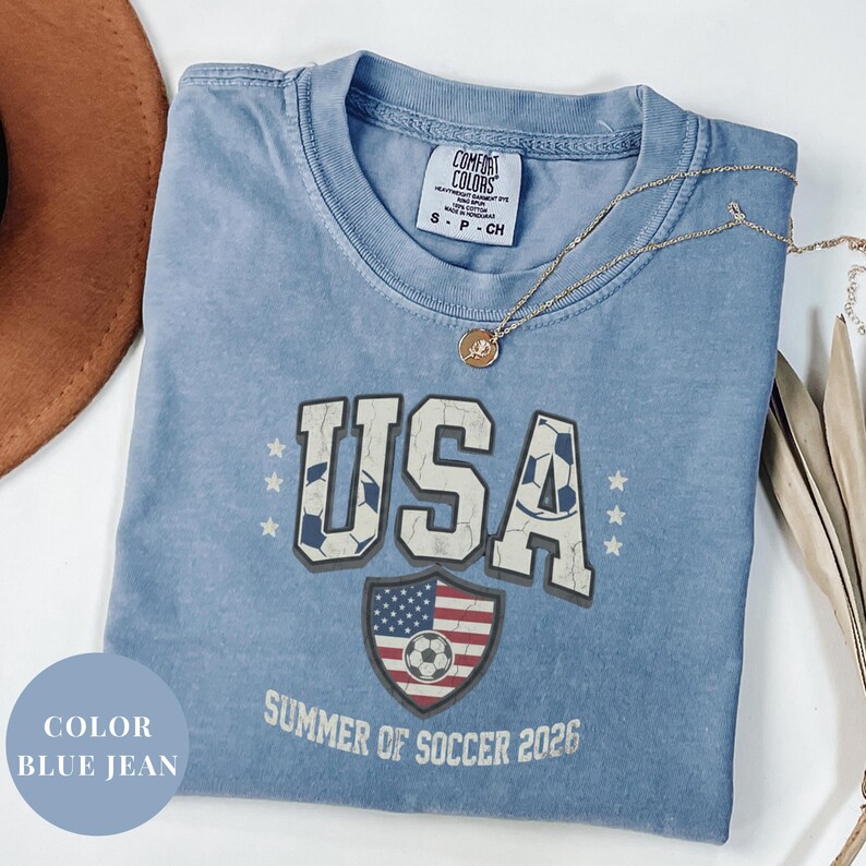 Blue Comfort Colors 1717 t-shirt with a vintage "USA Summer of Soccer 2026" graphic. Features bold varsity "USA" lettering with soccer ball patterns and a distressed American flag soccer shield. Patriotic apparel for World Cup 2026 fans.
