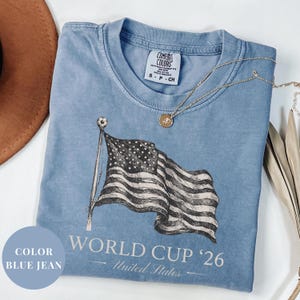 Blue Jean Comfort Colors 1717 t-shirt featuring a vintage hand-drawn sketch of a waving American flag. Includes "WORLD CUP '26" and "United States" text, with a subtle soccer ball detail on the flagpole. High-quality patriotic soccer apparel for the 2026 world tournament fans.