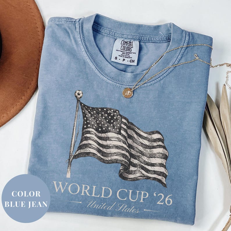 Blue Jean Comfort Colors 1717 t-shirt featuring a vintage hand-drawn sketch of a waving American flag. Includes "WORLD CUP '26" and "United States" text, with a subtle soccer ball detail on the flagpole. High-quality patriotic soccer apparel for the 2026 world tournament fans.