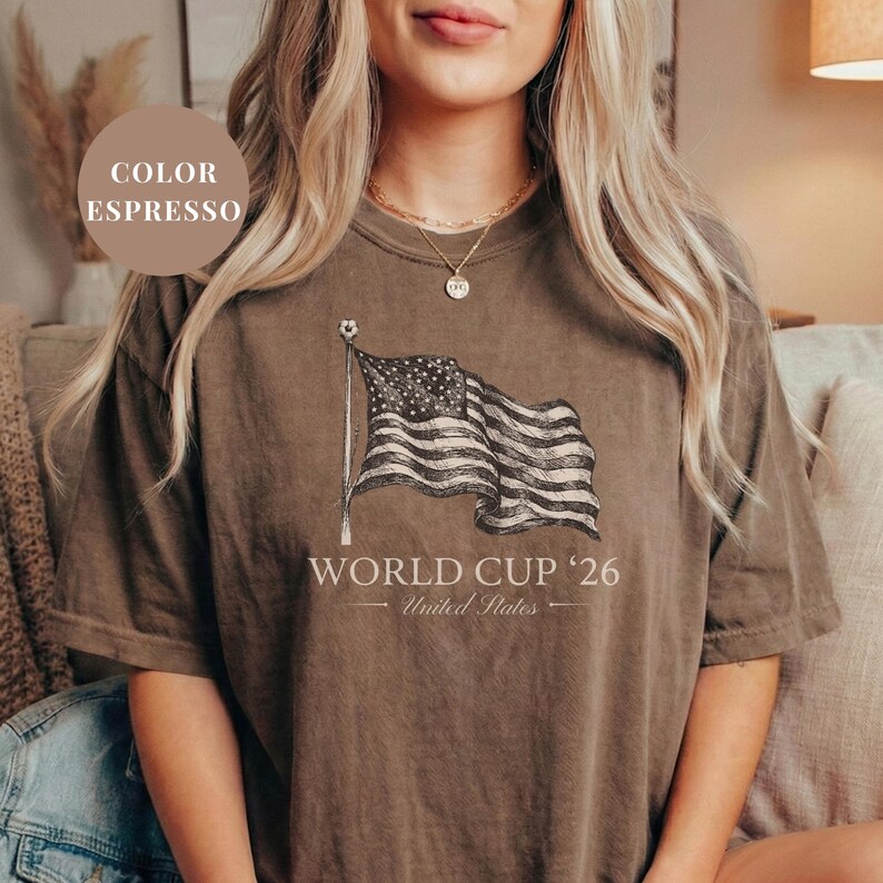 Espresso brown Comfort Colors 1717 t-shirt featuring a vintage hand-drawn sketch of a waving American flag. Includes "WORLD CUP '26" and "United States" text, with a subtle soccer ball detail on the flagpole. High-quality patriotic soccer apparel for the 2026 world tournament fans.