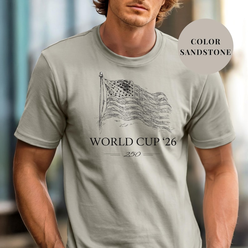 Sand Comfort Colors 1717 t-shirt featuring a minimalist vintage sketch of a waving American flag. Includes "WORLD CUP '26" and a stylized "250" text celebrating the USA Semiquincentennial. Premium patriotic sports apparel for United States soccer fans and 2026 host nation supporters.