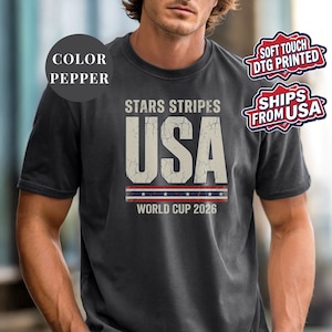 Pepper t-shirt featuring a distressed vintage "STARS STRIPES USA WORLD CUP 2026" graphic. The design includes weathered block lettering for "USA" and a patriotic red, white, and blue banner with stars. High-quality sports apparel for United States soccer fans and world tournament supporters.