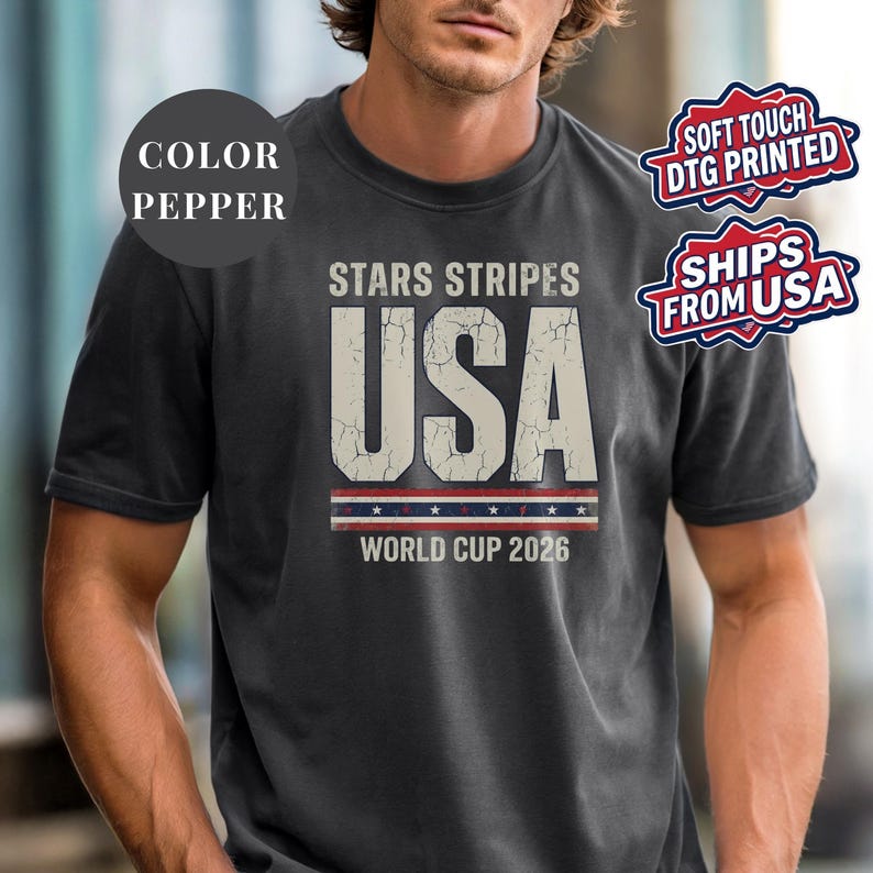 Pepper t-shirt featuring a distressed vintage "STARS STRIPES USA WORLD CUP 2026" graphic. The design includes weathered block lettering for "USA" and a patriotic red, white, and blue banner with stars. High-quality sports apparel for United States soccer fans and world tournament supporters.