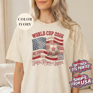 Ivory colored t-shirt featuring a vintage patchwork American flag design for World Cup 2026. The faux quilted graphic includes floral and gingham fabric textures, a stitched soccer ball, and "Stars, Stripes & Soccer" text. Unique patriotic sports apparel for women and USA soccer fans.